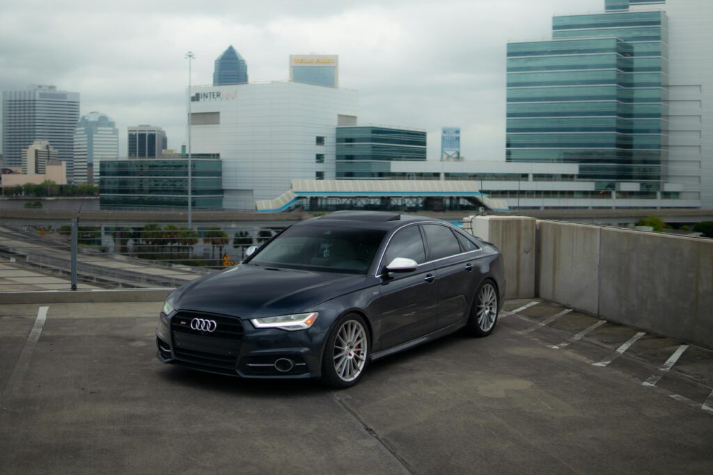 A sleek black Audi A6 sports car parked in a cityscape's urban parking lot, showcasing modern architecture.